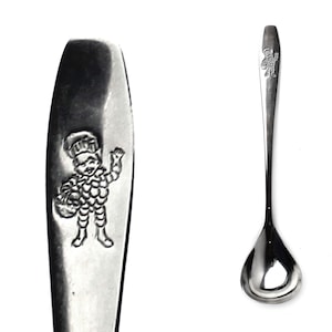 Op de afbeelding: Een zilveren lepel met een afgeronde kom en een lange steel. De steel heeft een reliëfbeeld van een chef-achtig figuur met een mand. De lepel is waarschijnlijk vintage en geschikt om te serveren of te eten.