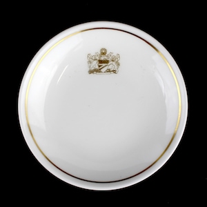 Op de afbeelding: Witte keramische schotel met een gouden rand en een gouden wapenschild in het midden. Het wapenschild toont een zon, twee vogels en een schild met de tekst "The Breakers" erop.