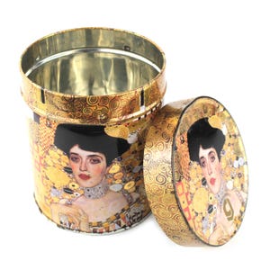 Op de afbeelding: Een ronde, open blikken doos met een deksel, met een portret van een vrouw met zwart haar en een decoratieve goud-gele achtergrond. Het ontwerp van de doos bevat een herhalend patroon van wervelingen en geometrische vormen, met een metalen interieur.