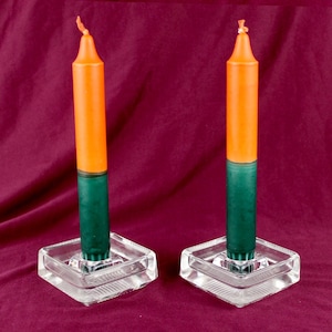 Può includere: Due candele arancioni e verdi in portacandele quadrati di vetro trasparente. Le candele sono divise in due colori, arancione in alto e verde in basso. I portacandele hanno un design strutturato. Lo sfondo è rosso scuro.
