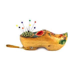 Könnte beinhalten: Ein holzschuhförmiges Nadelkissen mit einem grün-gelben Karokissen. Bunte Nadeln mit roten, gelben, blauen und grünen Köpfen sind in das Kissen gesteckt. Der Holzschuh hat bemalte Blumendetails und einen gelben Riemen.