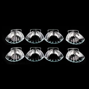 May include: Eight clear glass seashell-shaped dishes arranged in two rows of four against a black background. The dishes have a ridged texture and a small, flat base. The glass reflects light, creating highlights and shadows.