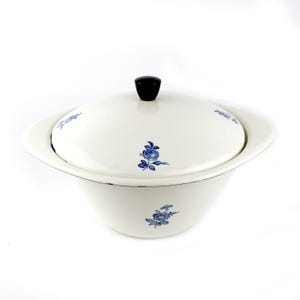 Può includere: Una zuppiera in ceramica bianca con coperchio. Il piatto presenta motivi floreali blu e un pomello nero sul coperchio. Il piatto ha un bordo largo e una forma arrotondata.