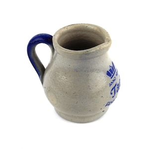 Könnte beinhalten: Eine kleine, graue Keramikkanne mit blauem Henkel und blauer Beschriftung. Die Beschriftung lautet "Anno 1780" und "Traubensaft".