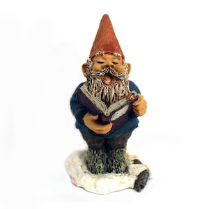 May include: A ceramic garden gnome figurine with a red pointed hat, blue jacket, and a long white beard. The gnome is holding a purple book and appears to be singing. The figurine is standing on a patch of white snow.