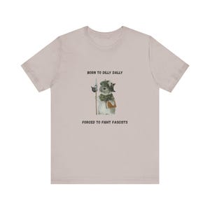 May include: A light pink t-shirt featuring a watercolor illustration of a rabbit dressed in medieval attire, holding a spear. The text above reads "BORN TO DILLY DALLY" and below, "FORCED TO FIGHT FASCISTS."
