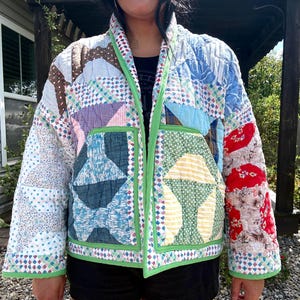May include: A quilted jacket constructed from various patchwork fabric squares in shades of blue, red, yellow, and white. The garment features a bright green trim along the front opening, collar, and cuffs, with two large square pockets on the front.