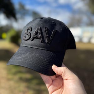 May include: A black baseball cap with the word "SAV" embroidered in black thread on the front.