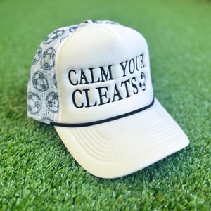 May include: White trucker hat with the text "CALM YOUR CLEATS" in black. The hat features a mesh back with a soccer ball pattern and a black rope detail along the brim. The hat is on a green surface.