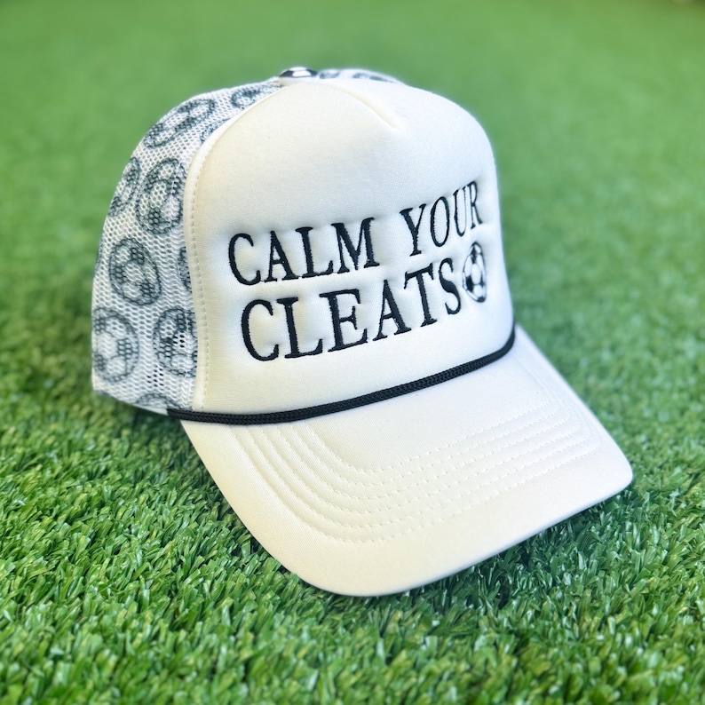 May include: White trucker hat with the text "CALM YOUR CLEATS" in black. The hat features a mesh back with a soccer ball pattern and a black rope detail along the brim. The hat is on a green surface.