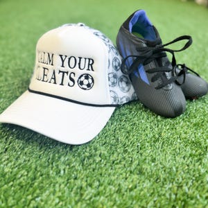 May include: A white trucker hat with the text "CALM YOUR CLEATS" and a soccer ball graphic. Next to the hat are black and blue soccer cleats with black laces, all on a green surface.