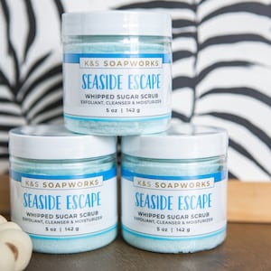 May include: Three white jars with blue lids and blue sugar scrub inside. The jars are labeled "K&S Soapworks Seaside Escape Whipped Sugar Scrub Exfoliant, Cleanser & Moisturizer 5 oz | 142 g".