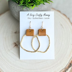 May include: A pair of gold-toned earrings with a brown leather accent. The earrings are displayed on a white card with the text "A Very Crafty Mommy Etsy - Quilted Bear - Locals".