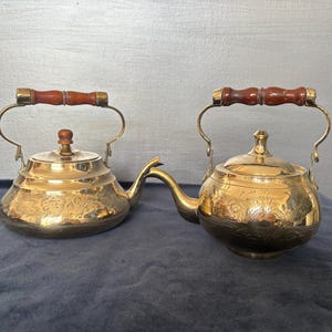May include: Two polished brass kettles featuring intricate floral engravings on the body, a rounded lid with a small knob, and a curved spout. Each kettle has a hinged metal handle with a dark brown, segmented wooden grip for handling.