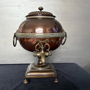 May include: A polished copper beverage dispenser with a spherical body, a removable lid, and a central spigot. The vessel features decorative silver-toned metal bands, side ring handles, and a square base supported by four small, rounded feet.