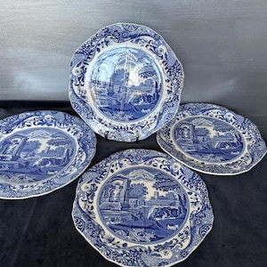 May include: A set of four round ceramic plates featuring a blue and white transferware pattern. Each plate displays a detailed landscape scene with architectural ruins, trees, and rolling hills, surrounded by a decorative floral border.