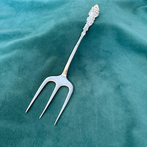 May include: Silver serving fork with three prongs, on a teal green velvet surface. The handle is ornate with a detailed floral design. A classic piece of silverware.
