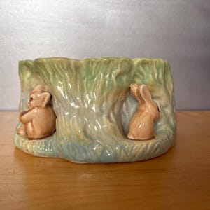 May include: A ceramic planter with a tree trunk design, featuring a seated monkey and a rabbit. The planter has a green and blue colour scheme, with the animals in a light brown hue. The planter is designed to hold plants.