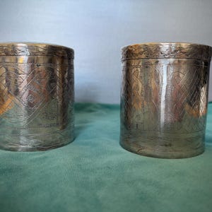 May include: Two silver-coloured, cylindrical containers with lids. The containers have intricate etched designs covering their surfaces. The background is a muted teal colour, providing a contrast to the metallic finish of the containers.