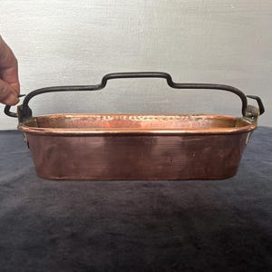 May include: A rectangular copper pan with a black metal handle. The pan has a shiny, reddish-brown surface and appears to be vintage. The handle is arched and attached to the sides of the pan. The pan is empty and sits on a dark blue surface.