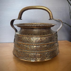 May include: An antique brass container with an arched handle and ornate floral patterns. The container has a round base and a lid. The handle and side handles are a lighter brass colour than the body. The container is likely used for storage or display.