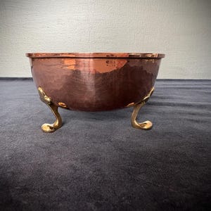 May include: A copper bowl with a hammered finish, supported by three decorative, gold-coloured feet. The bowl has a reddish-brown colour and a textured surface. The gold feet provide a contrast to the copper.