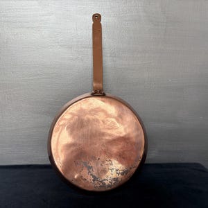 May include: A copper frying pan with a long, rectangular handle and a small hole for hanging. The pan's interior shows signs of use, with a dark patina. The pan is round, and the handle is attached with rivets.
