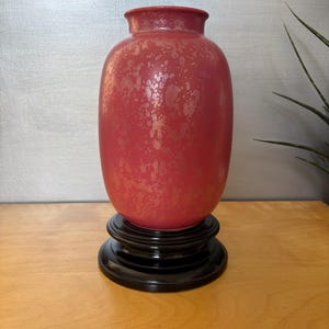 May include: A red vase with a speckled gold pattern, set on a black wooden stand. The vase has a rounded body and a slightly flared rim. The stand is a circular, tiered design.