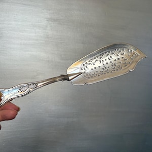 May include: Silver serving utensil with an ornate handle and a decorative, slotted head. The head has a delicate, openwork design. The utensil is held against a grey background.
