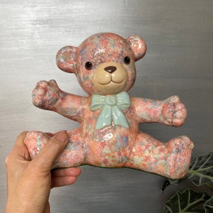 May include: A ceramic teddy bear figurine with a floral pattern in shades of pink, blue, and white. The bear is wearing a blue bow and has a light brown nose and eyes.