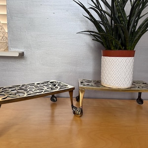 May include: Two brass plant stands with ornate, openwork designs. One stand is empty, while the other supports a white ceramic pot with a diamond pattern and a green plant. The stands have claw feet.
