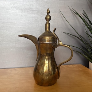 May include: A vintage brass coffee pot with a long spout, curved handle, and ornate lid. The pot has a polished finish and sits on a wooden surface. A decorative piece for serving coffee or tea.