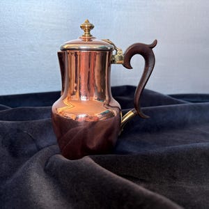 May include: A copper coffee pot with a dark wooden handle and lid. The pot has a shiny, reflective surface and a brass accent on the handle. The pot is sitting on a dark blue velvet surface.