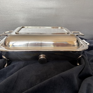 May include: A rectangular silver-coloured chafing dish with a polished metallic finish. The serving vessel features a removable lid with a flat top, two side handles, and four curved legs. A small, circular black knob is centred on the front side.