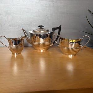 May include: A silver tea set comprising a teapot, creamer, and sugar bowl. The set features a ribbed design and is presented on a wooden surface. The teapot has a black handle and a decorative knob on the lid.