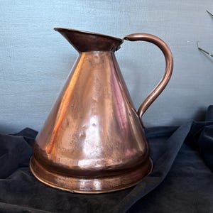 May include: A copper jug with a wide base and a curved handle. The jug has a conical shape and a dark brown rim. The copper has a warm, reddish-brown colour and a polished finish. The jug is likely used for serving water or other beverages.