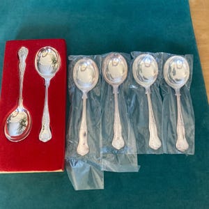 May include: Set of six silver soup spoons, five are in clear plastic packaging and one is in a red velvet box. The spoons have ornate handles.