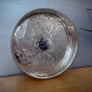 May include: A circular, silver-coloured tray featuring elaborate floral and scroll patterns. The tray has a detailed, rope-like edge and a decorative border. The centre of the tray has an engraved area. The tray is displayed on a wooden surface.