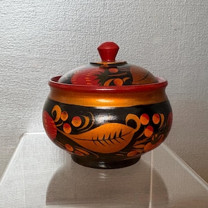 May include: A hand-painted wooden bowl with a lid. The bowl features a floral design in red, orange, and black. The lid has a red knob. The bowl is on a white surface.