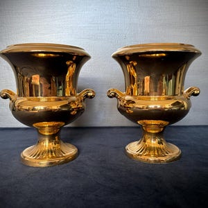 May include: A pair of gold-coloured urns with a dark interior. Each urn features a wide, flaring rim, a bulbous body, and a pedestal base. The urns have small, curved handles. The gold finish is shiny and reflective, typical of decorative items.