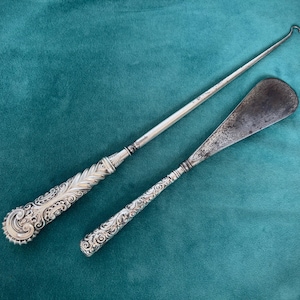 May include: Two antique silver tools with ornate handles on a teal surface. One is a long hook, the other a spatula-like instrument. The tools have detailed, engraved patterns. The background is teal.