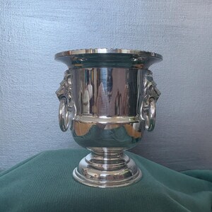 May include: A silver metal wine cooler with lion head handles. The cooler has a wide rim and a pedestal base. The cooler is highly reflective, and the background is a muted blue-grey.