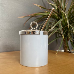 May include: A white ceramic sugar bowl with a silver lid and a silver spoon. The lid has a ring handle.