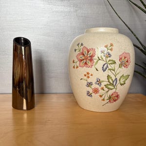 May include: A still life composition with two vases. A tall, bronze-coloured vase with a slanted top is next to a cream-coloured vase decorated with a floral pattern in pink, blue, and green. The vases are on a wooden surface.