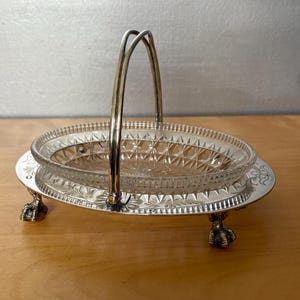 May include: An antique silver-plated serving dish with a clear glass insert. The dish has an oval shape with a decorative rim and is supported by claw feet. A curved handle rises from the dish.