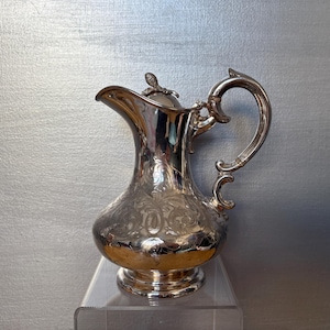 May include: A silver pitcher with an ornate design and a lid. The pitcher has a curved handle and a round body.