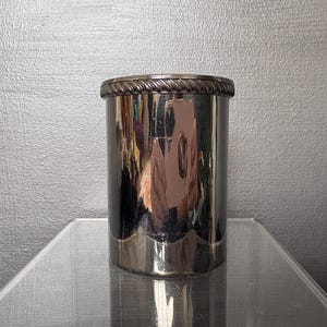 May include: A silver-coloured, cylindrical container with a decorative rope-like trim around the top. The container has a shiny, reflective surface and a scalloped base. It sits on a clear, reflective surface.