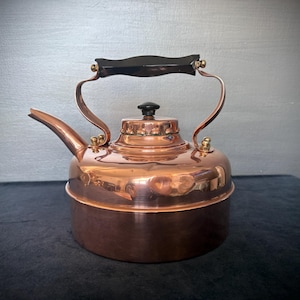 May include: A copper kettle with a black handle and lid knob. The kettle features a curved spout and a rounded body. The copper has a polished, reflective surface. The handle is attached with brass fittings. A classic kitchenware item.