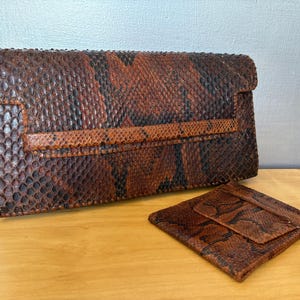May include: A brown and black snakeskin clutch and matching cardholder. The clutch has a rectangular shape with a flap closure and a strap detail. The cardholder is small and square with a flap. Both items have a textured surface.