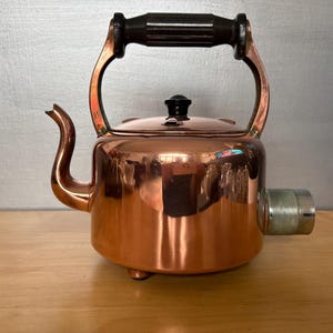 May include: A copper electric kettle with a black handle and lid knob. The kettle has a spout and a silver-coloured plug-in component. The kettle is on a wooden surface.
